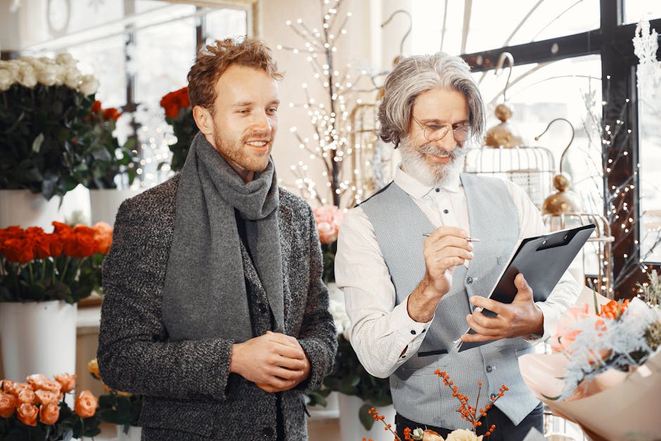 A man and an older male florist are inside a bright flower shop, surrounded by various fresh flowers including roses, carnations, and peonies in shades of red, pink, and white. The florist, wearing a 