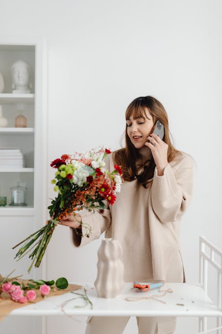 A young woman with long brown hair is smiling and speaking on a mobile phone while holding a vibrant floral bouquet. The bouquet features a mix of red, white, and pink flowers including roses and chry