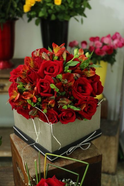 A vibrant floral arrangement featuring a bouquet of deep red roses, accented by smaller orange and yellow alstroemeria, with fresh green foliage interspersed throughout. The bouquet is arranged in a c