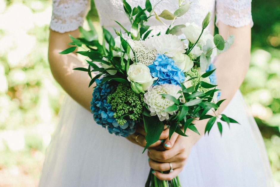 A person dressed in a white dress, holding a vibrant floral bouquet with both hands. The bouquet features large blue hydrangeas, white roses, and creamy white peonies, accented with green foliage and