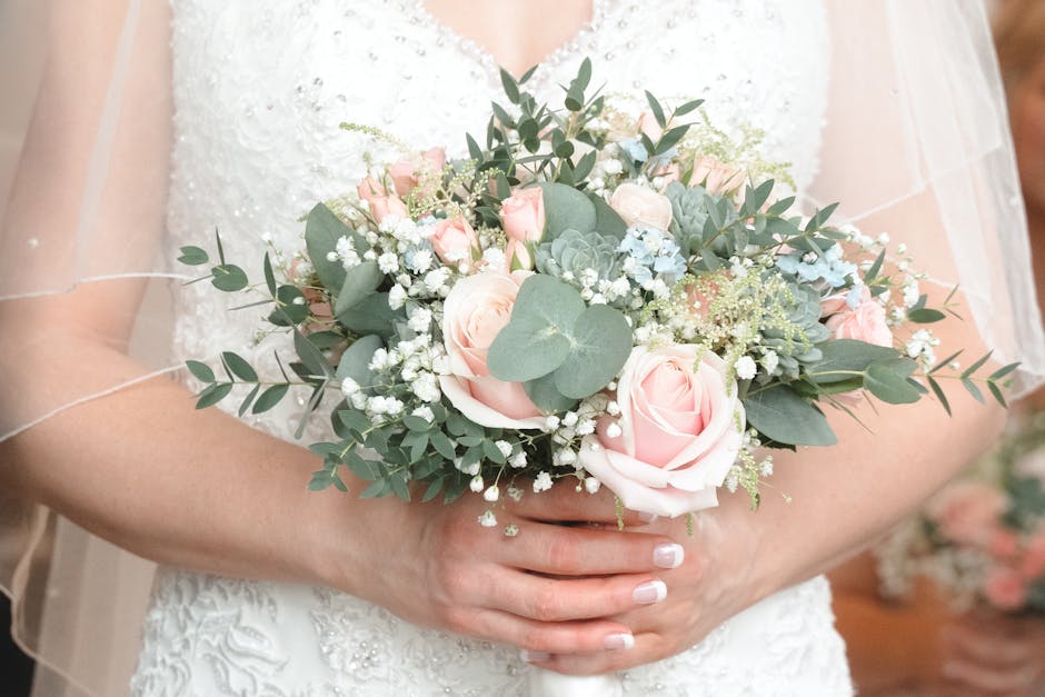 A bride wearing a white lace wedding dress and sheer veil holds a fresh floral bouquet consisting of pale pink roses, white baby's breath, green eucalyptus leaves, and succulents, arranged in a rounde