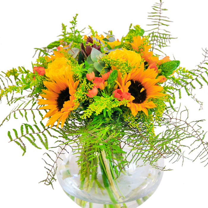 Infused with the vibrant energy of Wallington's community gardens and open green spaces, this lush bouquet is a visual and sensory feast. Sunflowers blaze at the heart of the arrangement, their wide, golden petals and deep brown centers imparting a sense of hope and brightness, much like the morning sun over Wallington Green. Surrounding them, intricately layered yellow garden roses bloom with elegant precision, the tight spirals of their petals opening in soft, golden-light reflections. Here and there, clusters of sparkling red Hypericum berries nestle among the blooms, their glossy presence elevating the entire palette and echoing the spirit of local summer fêtes. Trailing fern fronds and airy springs of solidago or goldenrod weave through the design, their lighter green hues and tiny blossoms suggesting the timeless beauty of nature in Carshalton's nearby valleys. Eucalyptus leaves add a touch of silvery coolness, and slender green stalks offer graceful structure, enhancing the bouquet's natural, hand-tied feel. The clear, rounded glass vase provides a modern touch-its watery depths revealing strong, healthy stems and anchoring the arrangement with transparency and authenticity. This bouquet, perfect for marking special milestones or as an uplifting gesture for loved ones, reflects the warmth and creativity found here in Wallington. Each element is chosen with care by your local florist, infusing the bouquet with the unique blend of joy and tradition that defines our neighbourhood.